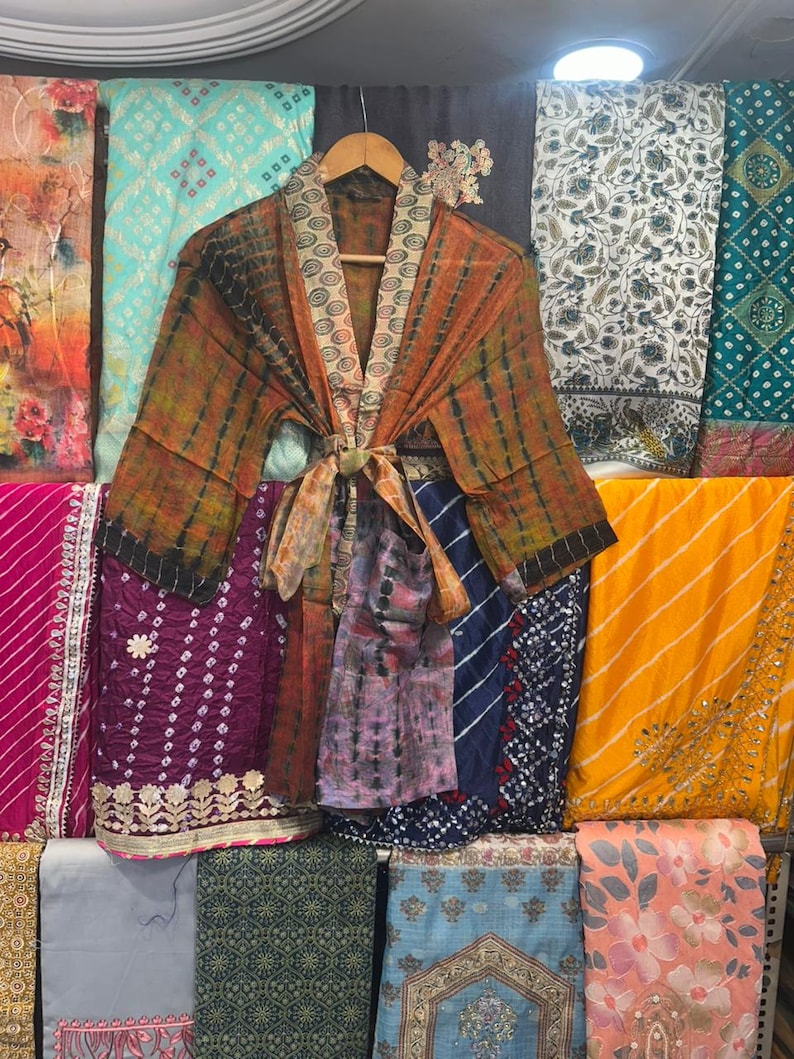 May include: A tie-dye kimono robe in shades of orange and brown, with a patterned collar and tie, displayed on a wooden hanger. The robe is surrounded by a variety of colourful fabrics with diverse patterns, including floral and geometric designs.