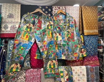May include: A two-piece pajama set with a colorful floral and figure print. The set includes a button-down shirt and pants, both hanging on a wooden hanger. The fabric has a light blue background with red, yellow, and green accents. The pajamas are displayed in a shop.