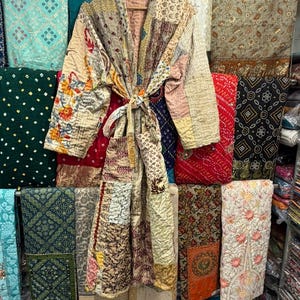 May include: A long-sleeved, knee-length robe featuring a patchwork design made from various quilted fabrics in muted tones of beige, cream, and soft pink, accented with floral embroidery on the left sleeve and a matching fabric tie belt at the waist.