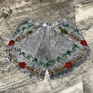 May include: Gray shorts with an embroidered floral design in red, green, and gold. The shorts have a drawstring waist and a textured fabric. The floral embroidery is along the edges of the shorts.