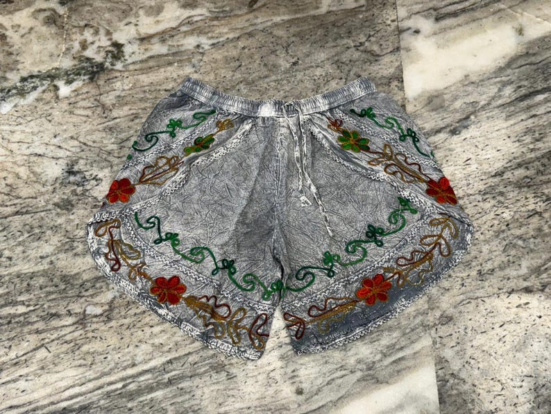 May include: Gray shorts with an embroidered floral design in red, green, and gold. The shorts have a drawstring waist and a textured fabric. The floral embroidery is along the edges of the shorts.