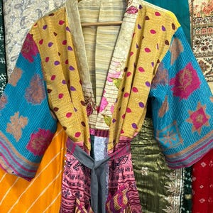 May include: A colorful, patchwork kimono robe with a tie belt. The robe features a mix of patterns and colors, including yellow, blue, pink, and floral designs. It hangs on a wooden hanger, showcasing its unique, bohemian style.