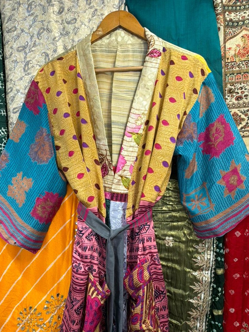 May include: A colorful, patchwork kimono robe with a tie belt. The robe features a mix of patterns and colors, including yellow, blue, pink, and floral designs. It hangs on a wooden hanger, showcasing its unique, bohemian style.