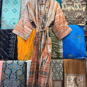 May include: A long-sleeved kimono robe featuring a tie-dye pattern in shades of peach, orange, and black. The garment includes a matching fabric belt at the waist, wide sleeves, and a front opening, displayed on a wooden hanger.