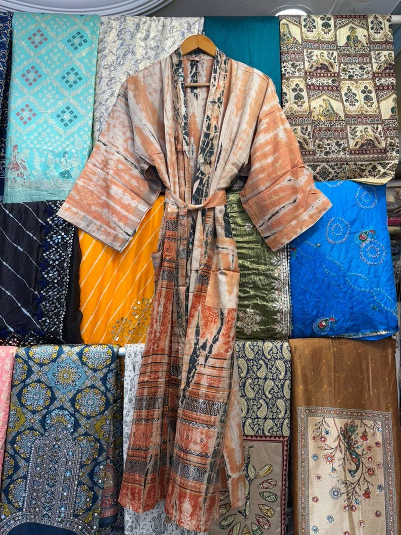 May include: A long-sleeved kimono robe featuring a tie-dye pattern in shades of peach, orange, and black. The garment includes a matching fabric belt at the waist, wide sleeves, and a front opening, displayed on a wooden hanger.