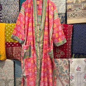 May include: A long-sleeved kimono-style robe in a vibrant pink hue, featuring a repeating pattern of yellow and white floral motifs. The garment includes a light green trim along the front opening and cuffs, with two front pockets and a matching belt.