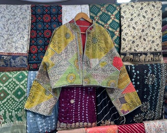 Op de afbeelding: Een patchwork jasje met gele, groene en rode stoffen. Het jasje heeft een gewatteerd ontwerp en hangt aan een houten hanger. De achtergrond bestaat uit diverse stoffen met patronen.