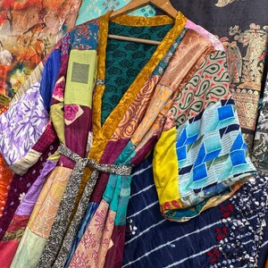 May include: A patchwork kimono robe with a variety of colorful fabric patterns, including paisley, floral, and geometric designs. The robe has a gold trim and a matching belt. It is hanging on a wooden hanger.