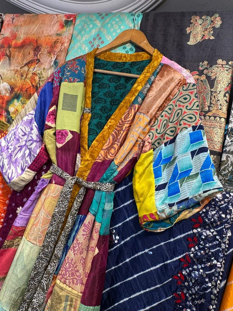 May include: A patchwork kimono robe with a variety of colorful fabric patterns, including paisley, floral, and geometric designs. The robe has a gold trim and a matching belt. It is hanging on a wooden hanger.