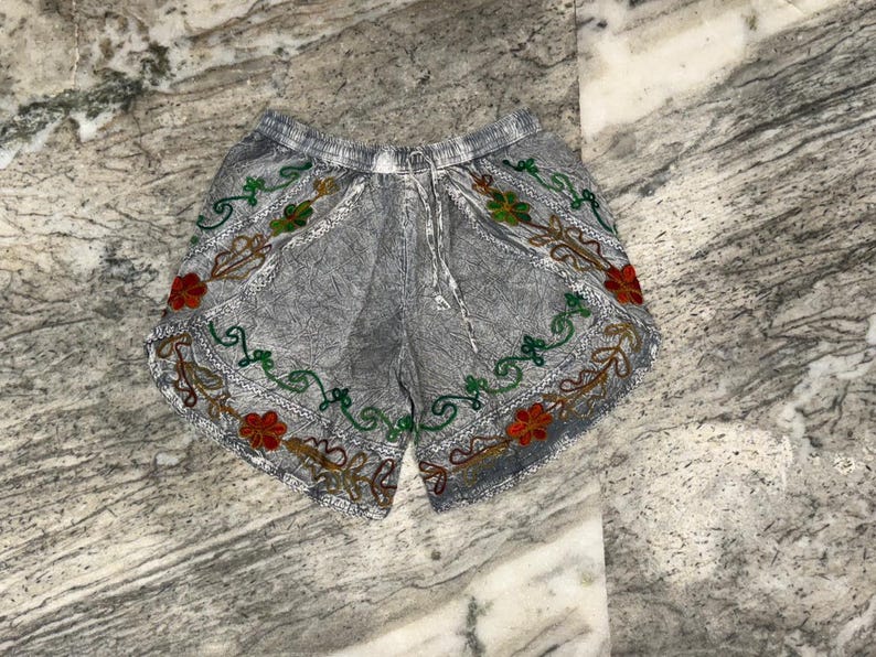 May include: Gray shorts with an embroidered floral design in red, green, and brown. The shorts have a drawstring waist and are made of a textured fabric. The floral embroidery is along the bottom edge of the shorts.