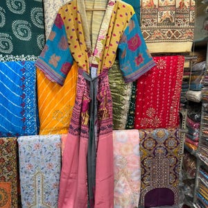 May include: A colorful, patterned kimono robe with a tie belt hangs on a wooden hanger. The robe features blue, yellow, pink, and patterned fabric. Various textiles in different colors and patterns are visible in the background.