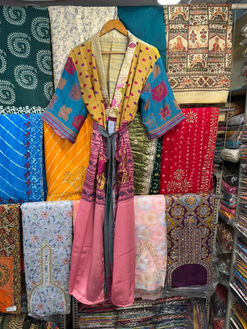 May include: A colorful, patterned kimono robe with a tie belt hangs on a wooden hanger. The robe features blue, yellow, pink, and patterned fabric. Various textiles in different colors and patterns are visible in the background.