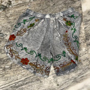 May include: Gray shorts with an elastic waistband and drawstring. The shorts are decorated with colorful embroidered floral and vine patterns in red, green, and gold. The fabric has a textured appearance.
