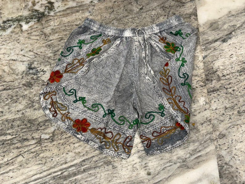 May include: Gray shorts with an elastic waistband and drawstring. The shorts are decorated with colorful embroidered floral and vine patterns in red, green, and gold. The fabric has a textured appearance.