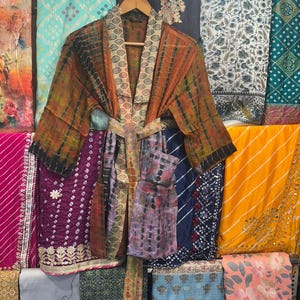 May include: A tie-dye patterned kimono robe in shades of orange, brown, and green hangs on a wooden hanger. The robe features a patterned collar and a matching tie belt. Various colourful fabrics are visible in the background.