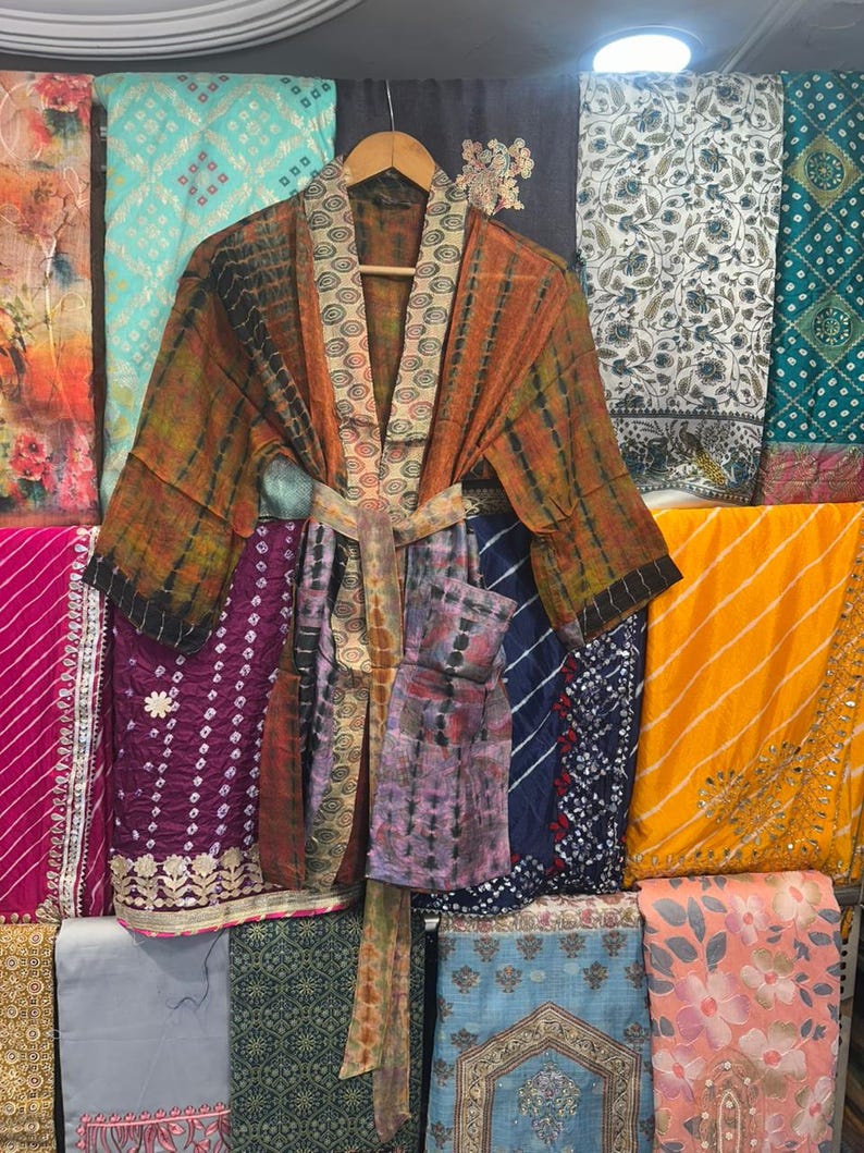 May include: A tie-dye patterned kimono robe in shades of orange, brown, and green hangs on a wooden hanger. The robe features a patterned collar and a matching tie belt. Various colourful fabrics are visible in the background.