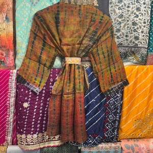 May include: A tie-dye patterned robe in shades of orange, brown, and black hangs on a wooden hanger. The robe is belted at the waist with a decorative band. Various colourful fabrics are visible in the background.