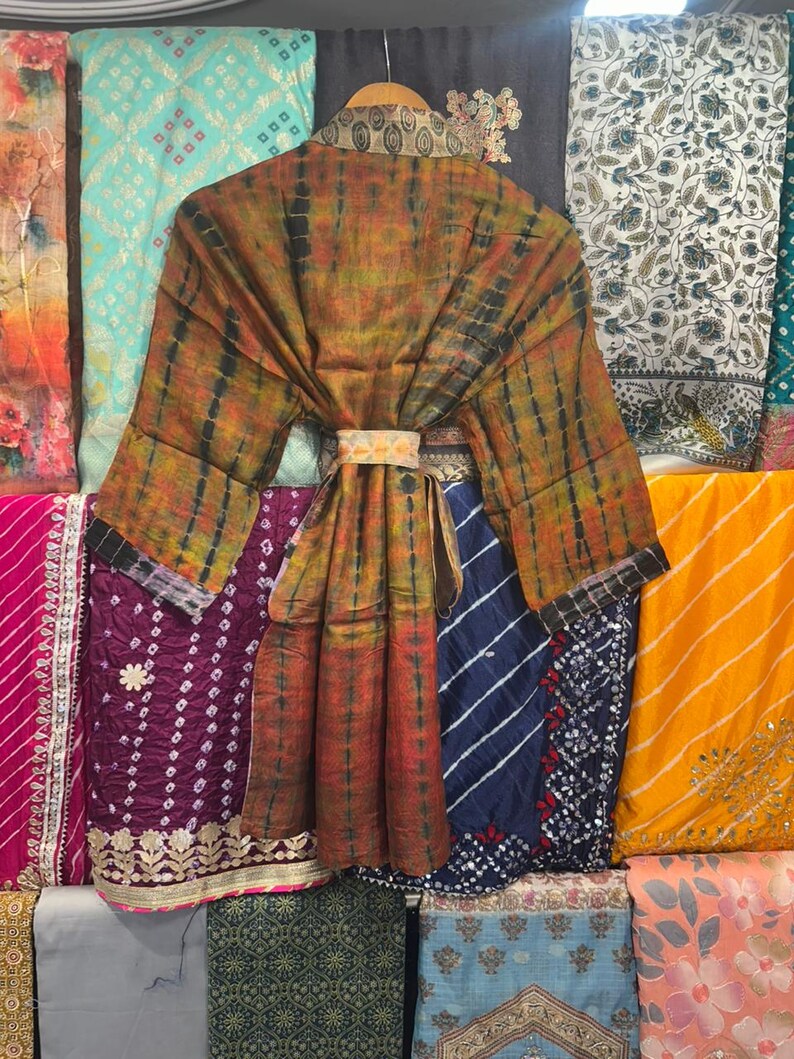 May include: A tie-dye patterned robe in shades of orange, brown, and black hangs on a wooden hanger. The robe is belted at the waist with a decorative band. Various colourful fabrics are visible in the background.