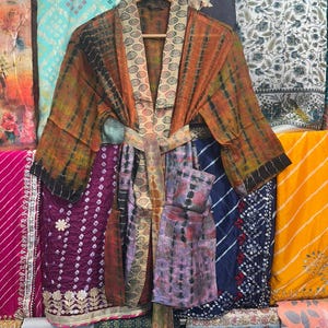 May include: A tie-dye patterned kimono robe in shades of orange, brown, and black, with a matching belt. The robe is displayed on a wooden hanger, set against a backdrop of patterned fabric panels.