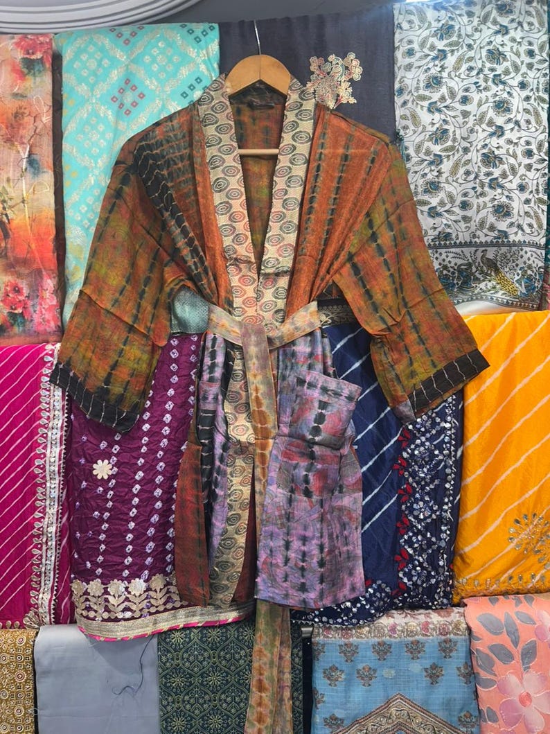 May include: A tie-dye patterned kimono robe in shades of orange, brown, and black, with a matching belt. The robe is displayed on a wooden hanger, set against a backdrop of patterned fabric panels.