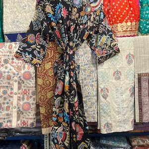May include: A black robe with a colourful floral design in blue, pink, yellow, and green. The robe is tied at the waist and hangs on a wooden hanger. Other patterned fabrics are visible in the background.