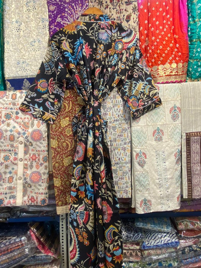 May include: A black robe with a colourful floral design in blue, pink, yellow, and green. The robe is tied at the waist and hangs on a wooden hanger. Other patterned fabrics are visible in the background.