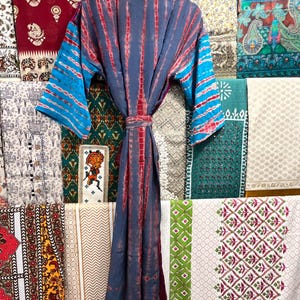 May include: A blue and red tie-dye kimono robe with a long sleeve and a sash. The robe is hanging on a rack in front of a wall of colorful patterned fabrics.