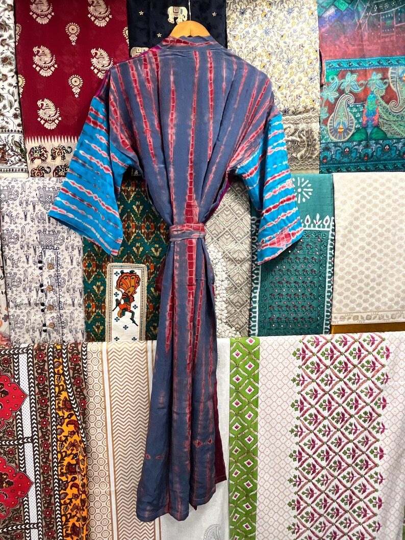 May include: A blue and red tie-dye kimono robe with a long sleeve and a sash. The robe is hanging on a rack in front of a wall of colorful patterned fabrics.