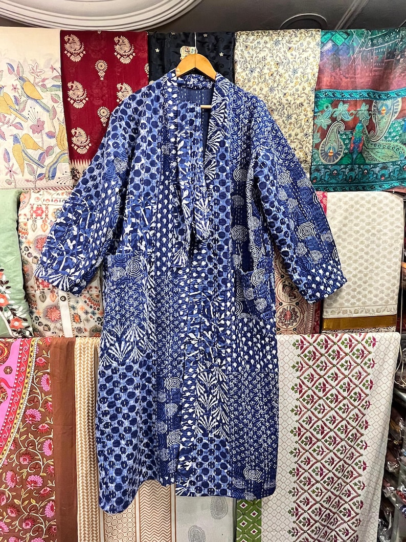 Op de afbeelding: Een blauwe en witte geruite gequilte badjas met lange mouwen en een striksluiting. De badjas hangt aan een hanger voor een muur van kleurrijk stof.