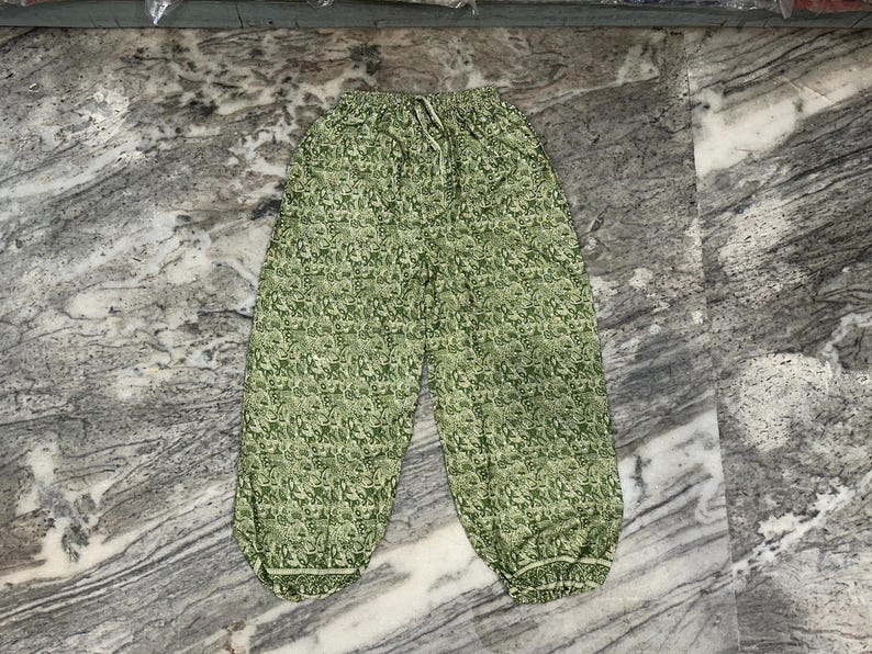 May include: Green patterned trousers with a drawstring waist and elasticated cuffs. The trousers feature a repeating floral design in shades of green. The fabric appears lightweight and suitable for casual wear.