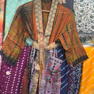 May include: A lightweight kimono robe featuring a tie-dye pattern in shades of orange, green, and black. The garment includes a wide collar and a matching tie belt with a circular geometric print, along with long sleeves and a front pocket.