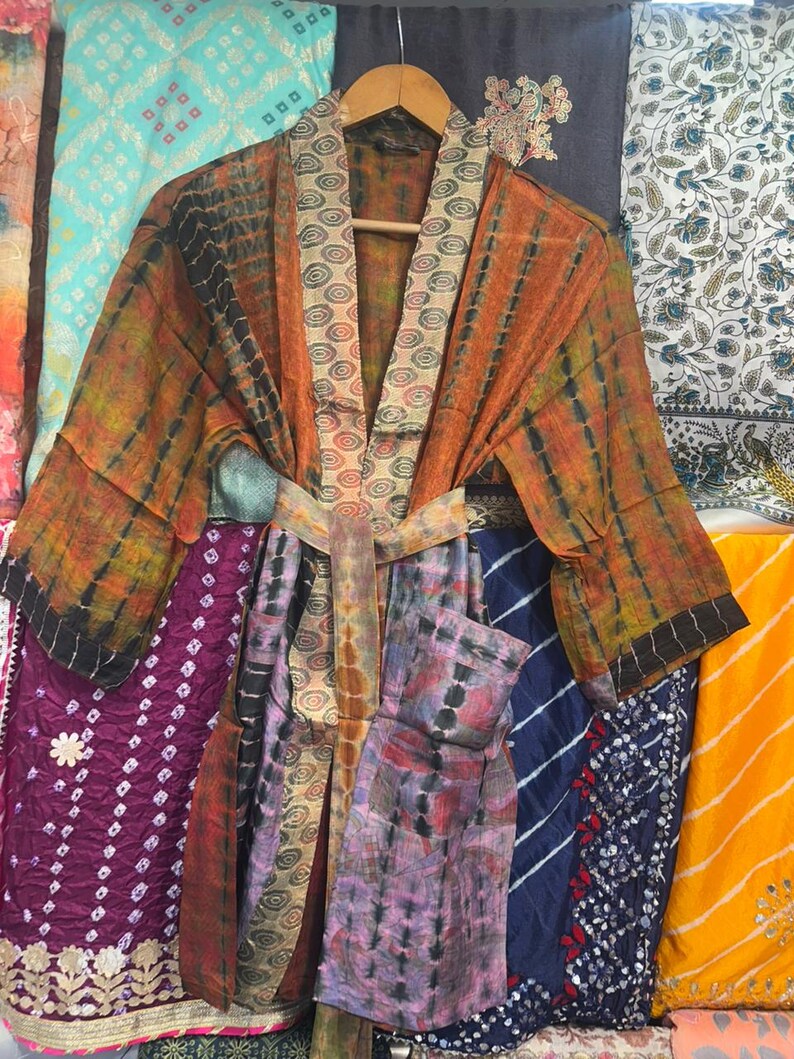 May include: A lightweight kimono robe featuring a tie-dye pattern in shades of orange, green, and black. The garment includes a wide collar and a matching tie belt with a circular geometric print, along with long sleeves and a front pocket.