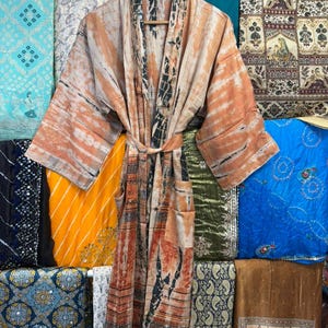 May include: A long-sleeved kimono robe featuring a tie-dye pattern in shades of tan, orange, and black. The garment includes a matching fabric belt at the waist, wide sleeves, and a vertical panel design along the front opening and hemline.