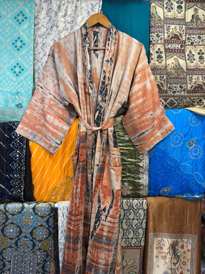 May include: A long-sleeved kimono robe featuring a tie-dye pattern in shades of tan, orange, and black. The garment includes a matching fabric belt at the waist, wide sleeves, and a vertical panel design along the front opening and hemline.