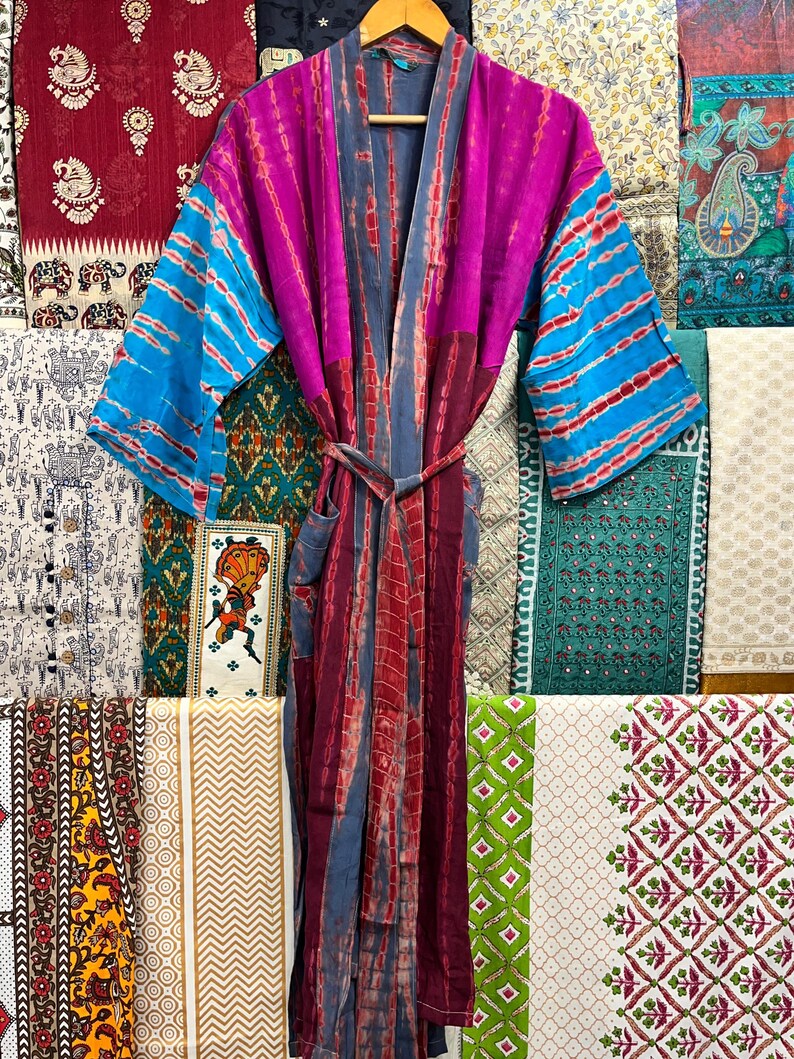 May include: A long, purple, blue, and red tie-dye robe with a sash. The robe is made of a soft, silky fabric and has a relaxed fit. The robe is hanging on a wooden hanger in front of a wall of colorful fabric.