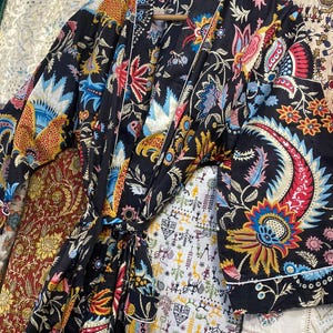 May include: A black patterned robe featuring a colourful floral design in red, blue, yellow, and orange. The robe has a white trim and a matching belt. It is displayed on a wooden hanger.