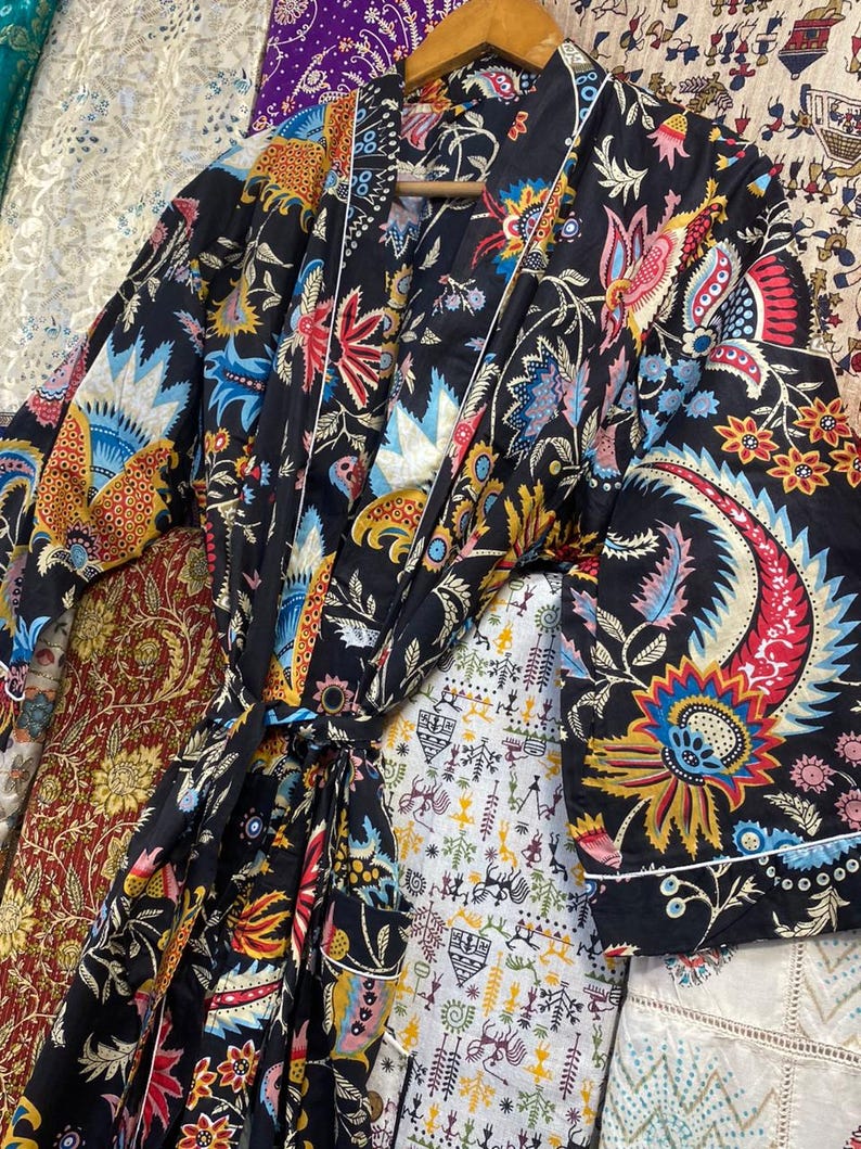 May include: A black patterned robe featuring a colourful floral design in red, blue, yellow, and orange. The robe has a white trim and a matching belt. It is displayed on a wooden hanger.
