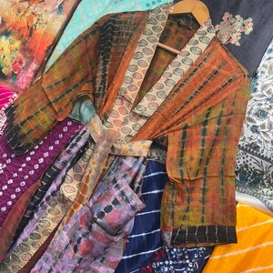 May include: A tie-dye kimono robe in orange and black, with a patterned collar and belt. The robe hangs on a wooden hanger, surrounded by colourful fabrics with diverse patterns and colours, including purple, blue, and yellow.