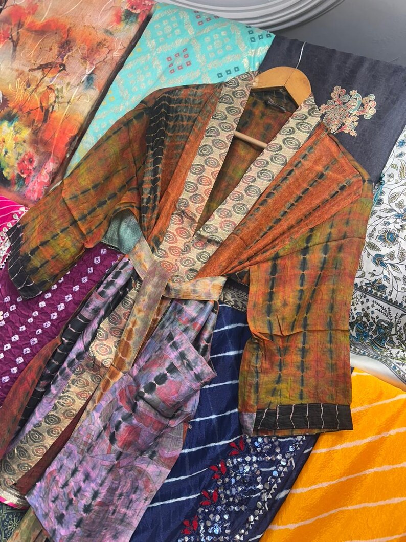 May include: A tie-dye kimono robe in orange and black, with a patterned collar and belt. The robe hangs on a wooden hanger, surrounded by colourful fabrics with diverse patterns and colours, including purple, blue, and yellow.
