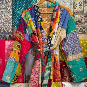 May include: A long-sleeved robe crafted from a patchwork of multicoloured cotton fabrics featuring various floral, geometric, and abstract patterns. The garment includes a matching tie belt and visible white running stitch quilting throughout.