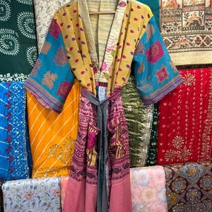 May include: A colorful, patterned kimono robe with a matching belt hangs on a wooden hanger. The robe features a mix of yellow, blue, and pink fabrics with floral and geometric designs. The background showcases various textiles.