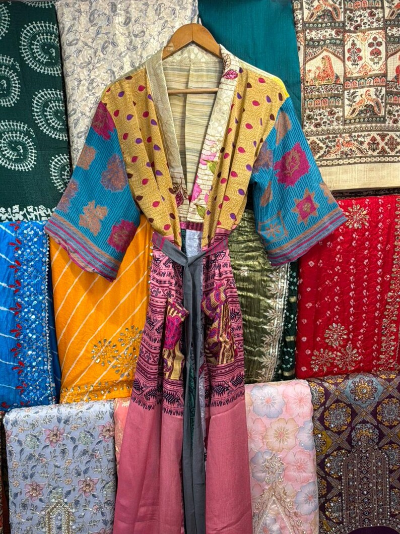 May include: A colorful, patterned kimono robe with a matching belt hangs on a wooden hanger. The robe features a mix of yellow, blue, and pink fabrics with floral and geometric designs. The background showcases various textiles.