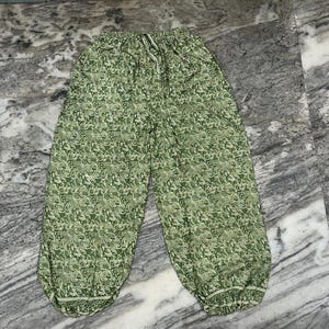 May include: Green patterned trousers with a drawstring waist. The trousers feature a repeating floral design in shades of green and white. The trousers have a relaxed fit and are made of a lightweight fabric.