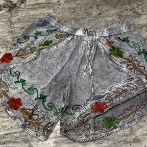 May include: Gray shorts with an elastic waistband and drawstring. The shorts feature colorful embroidered floral and vine designs in red, green, and gold along the edges. The fabric has a textured appearance.