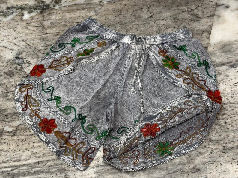 May include: Gray shorts with an elastic waistband and drawstring. The shorts feature colorful embroidered floral and vine designs in red, green, and gold along the edges. The fabric has a textured appearance.