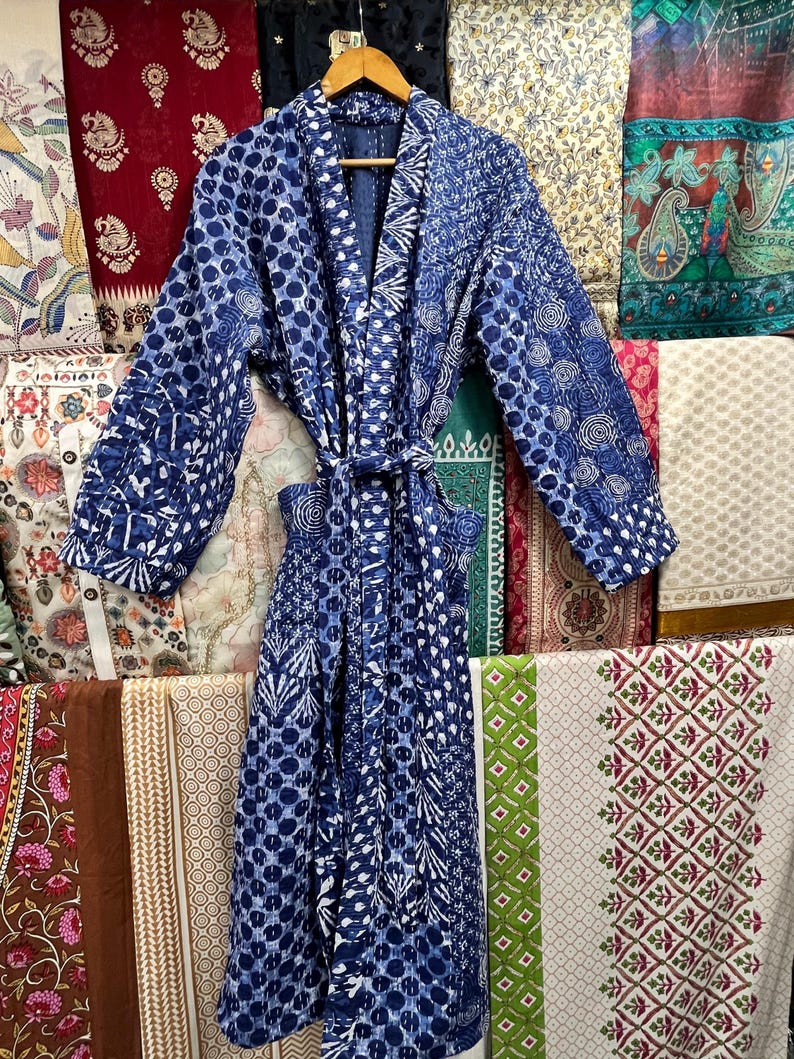 Op de afbeelding: Een blauwe en witte gequilte badjas met een strikceintuur. De badjas hangt aan een hanger voor een muur van kleurrijk stof.