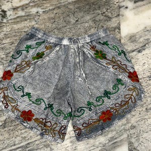 May include: Gray shorts with an elastic waistband and drawstring. The shorts are decorated with embroidered floral patterns in red, green, and gold. The fabric has a textured appearance.