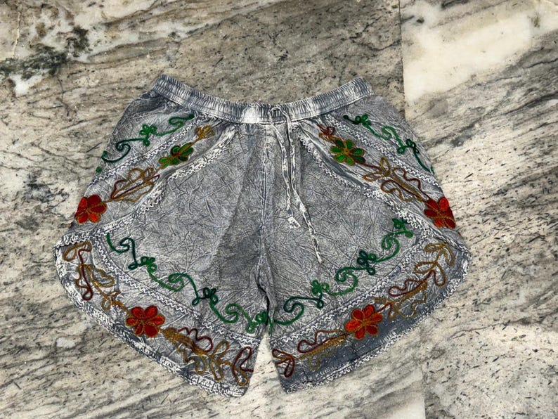 May include: Gray shorts with an elastic waistband and drawstring. The shorts are decorated with embroidered floral patterns in red, green, and gold. The fabric has a textured appearance.