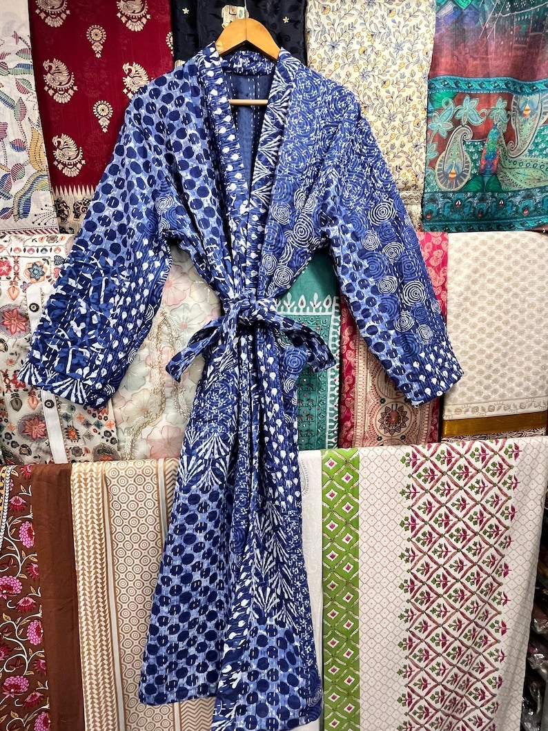 Op de afbeelding: Een blauwe en witte katoenen badjas met een strikceintuur. De badjas is gemaakt van een patchwork van verschillende stoffen en heeft een losse pasvorm.