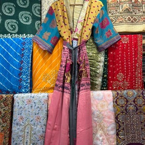 May include: A colorful, patterned kimono robe with a gray tie, hanging on a wooden hanger. The robe features a mix of yellow, pink, and blue fabrics. The background displays various patterned fabrics in different colors.